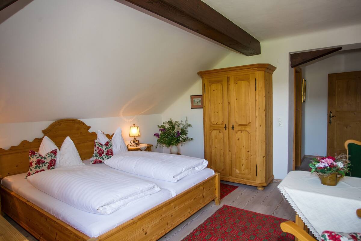 A bedroom with a wooden bed, a nightstand with a lamp, a wooden wardrobe, and a table with a vase of flowers.