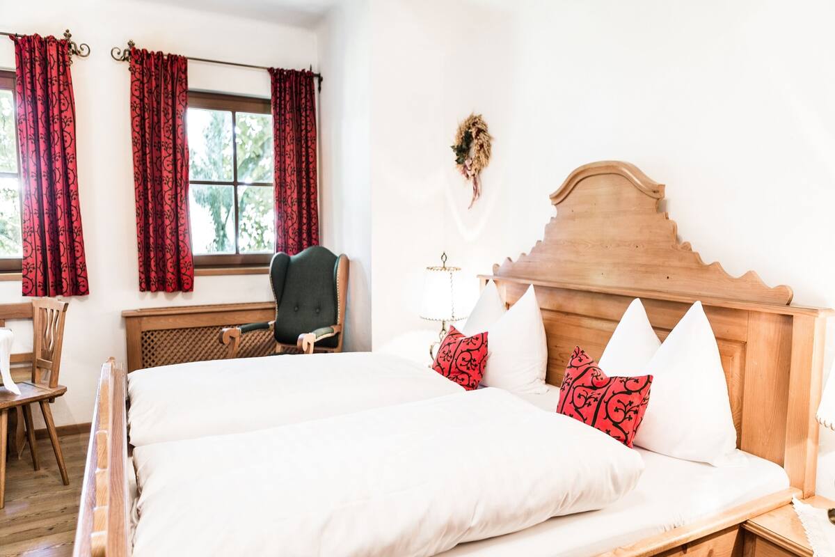 A bedroom with a wooden bed, red patterned curtains, a wooden chair, and a small desk with a lamp.
