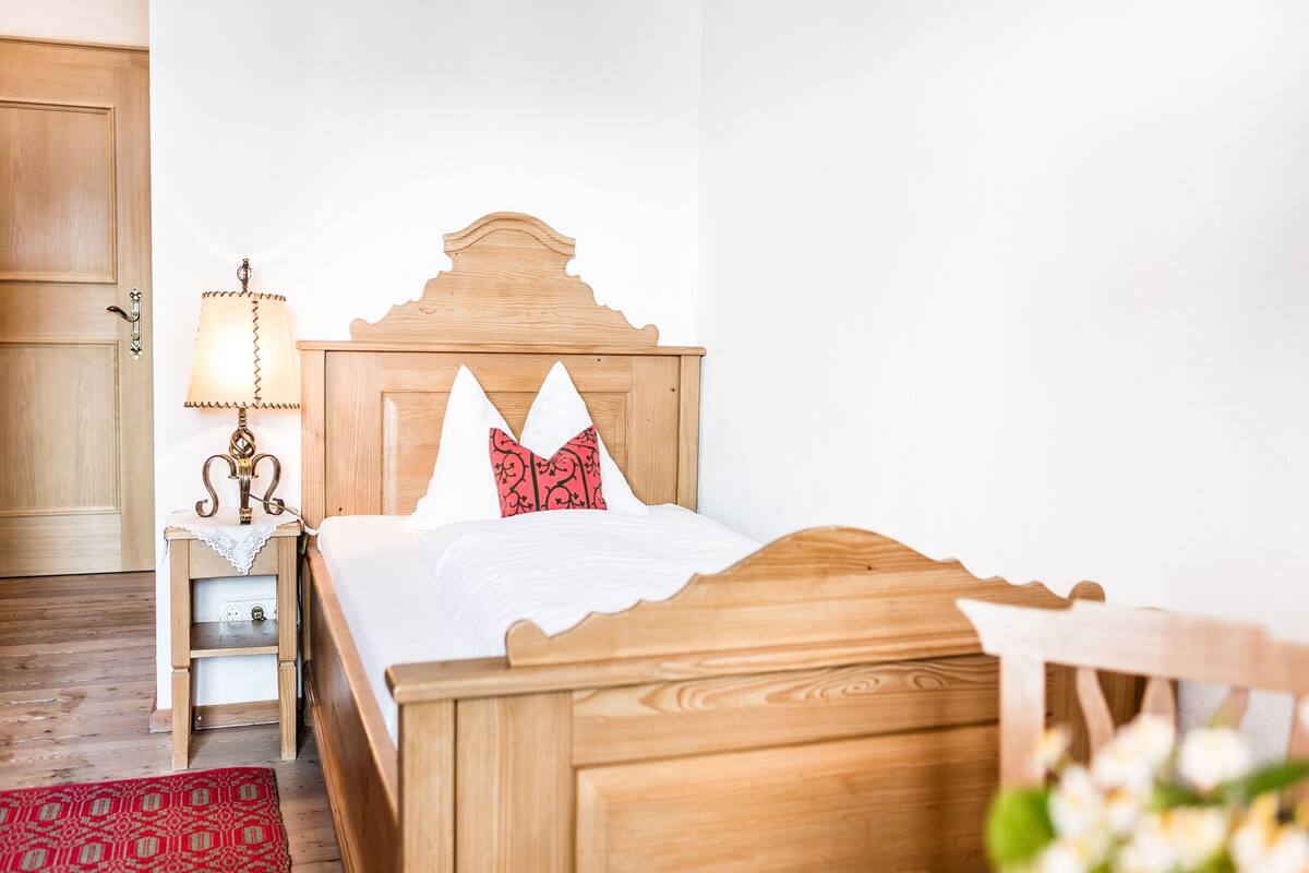 A wooden bed with a white mattress and two pillows, a bedside table with a lamp, and a red patterned rug.