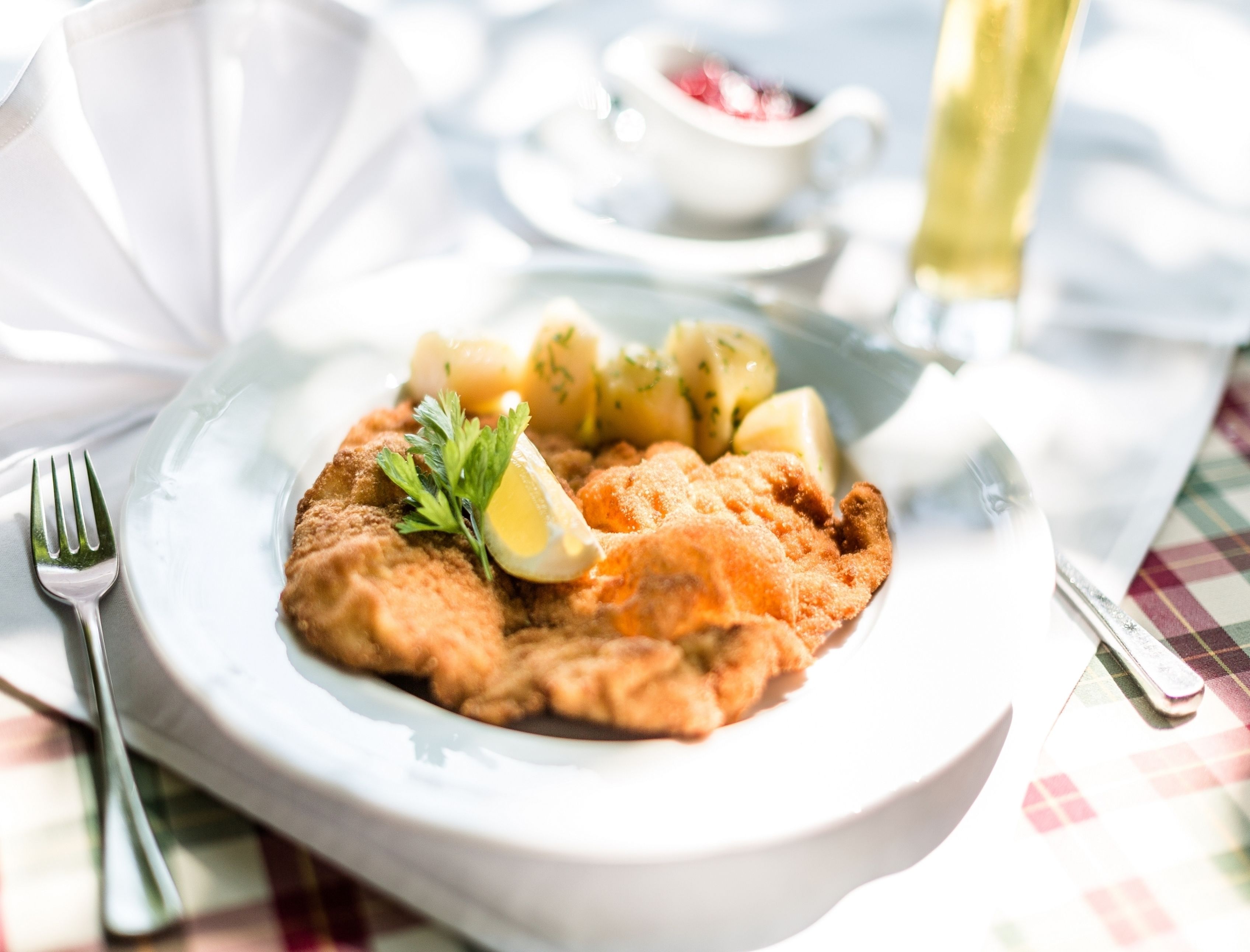 A plate with a breaded cutlet, potatoes, and a lemon wedge, accompanied by a fork and knife on a table with a checkered tablecloth.