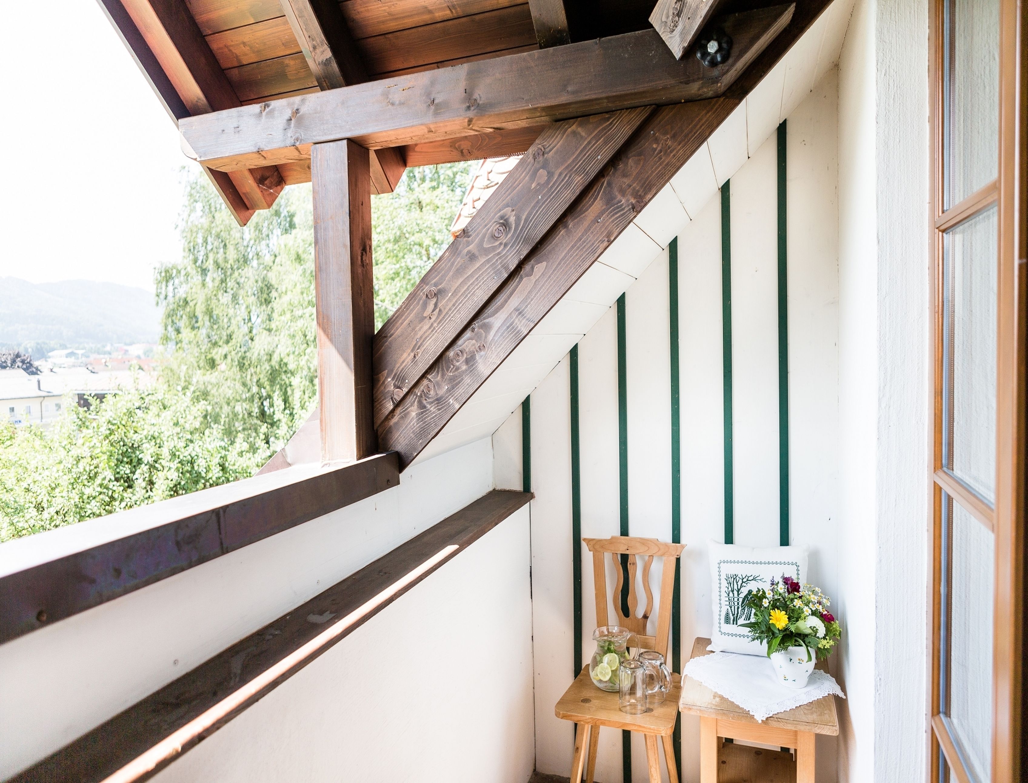 A balcony with wooden beams, a chair, a small table with a vase of flowers, and a view of greenery and buildings.