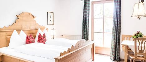 A wooden bed with white and red pillows, a wooden nightstand, a table with a chair, and a window with curtains.