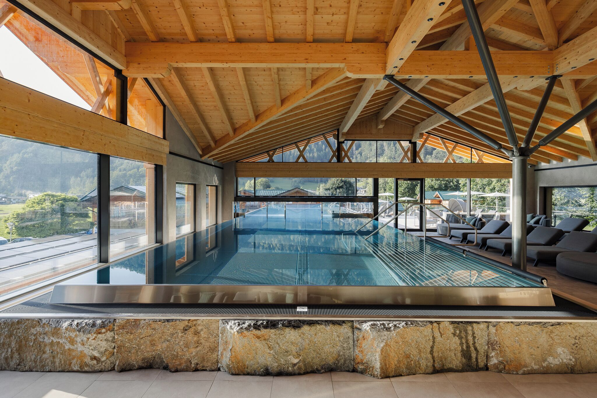 A modern indoor swimming pool with a view of the mountains, wooden ceiling beams, and lounge area with chairs.