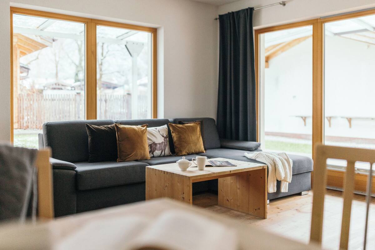 Een gezellige woonkamer met een donkergrijze bank, een houten salontafel en grote ramen met uitzicht op de achtertuin.