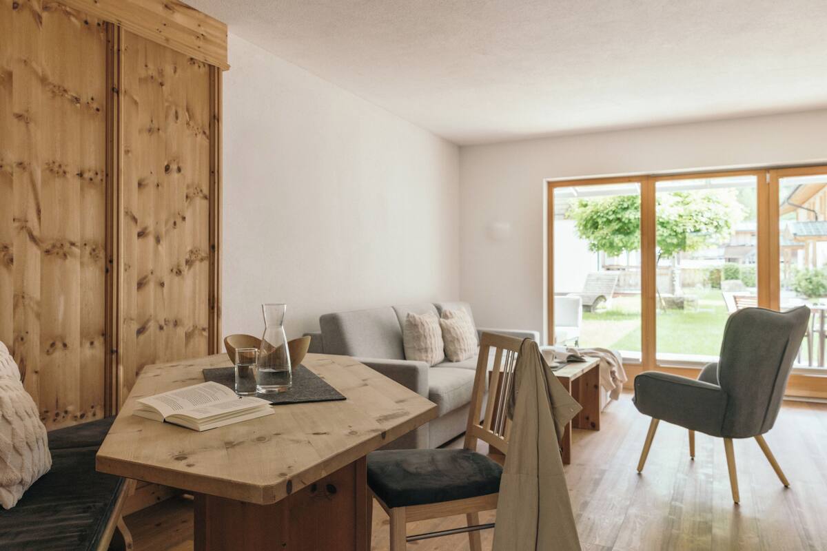 Een moderne woonkamer met een houten eettafel, een grijze bank en een groot raam met uitzicht op de omgeving.