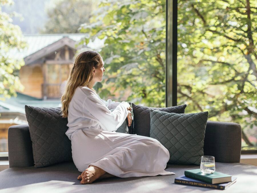 Uma pessoa sentada num sofá com um livro e um copo de água, num quarto com uma janela grande e vista para o verde lá fora.