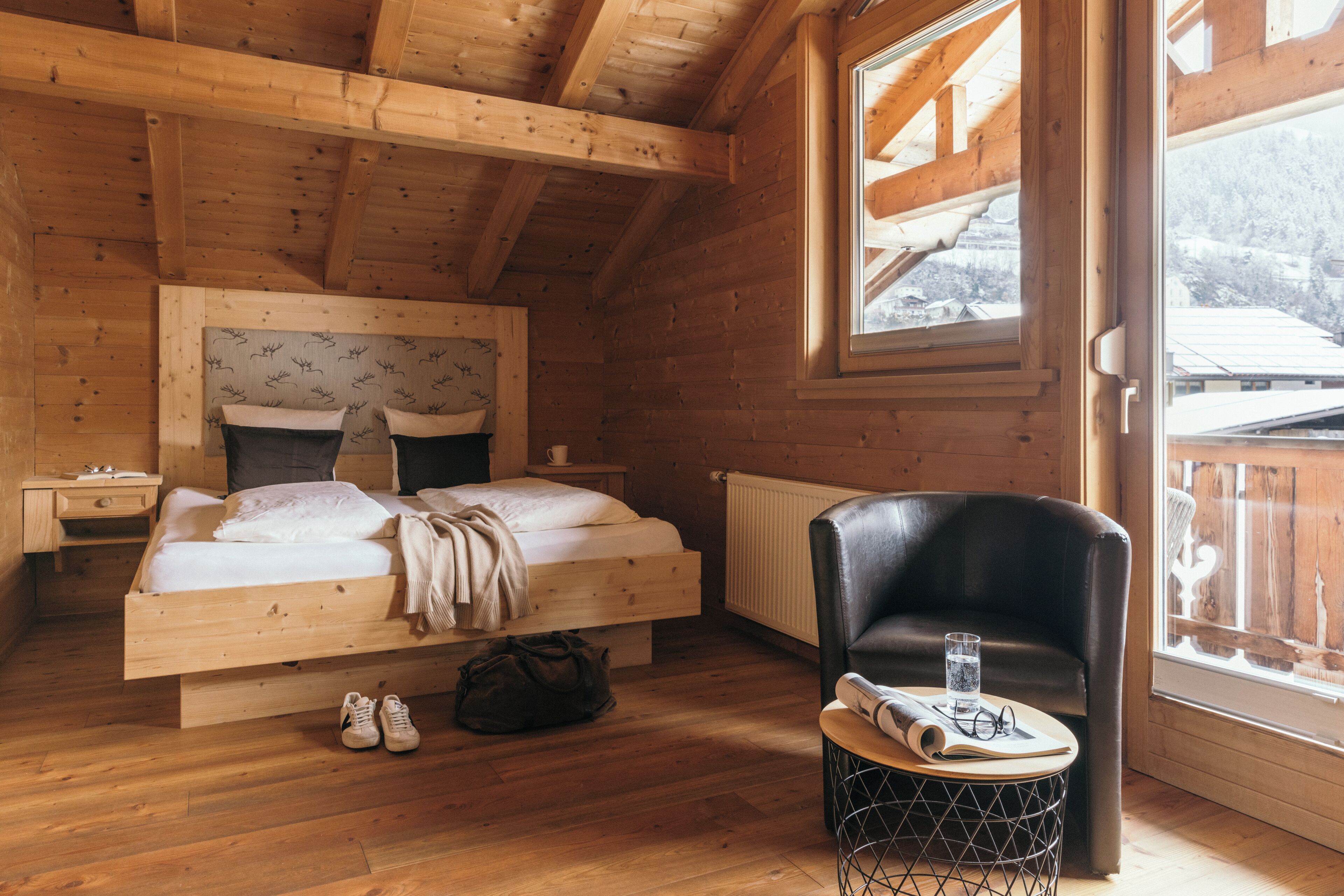 A wooden cabin bedroom with a bed, a chair, a small table, and a window with a view of a snowy landscape.