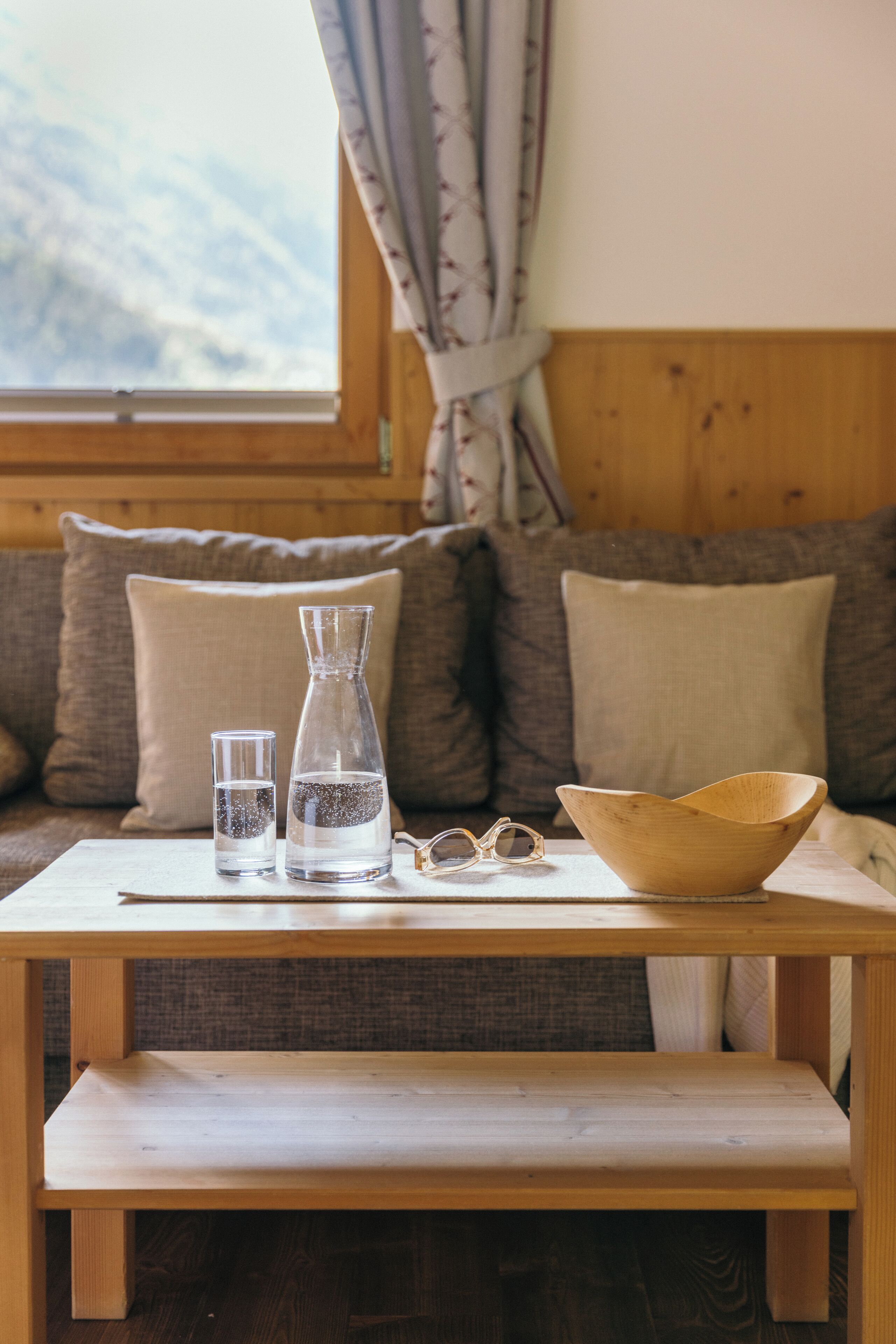 A wooden table with a glass pitcher, a glass, and a wooden bowl on it, placed in front of a sofa with cushions.