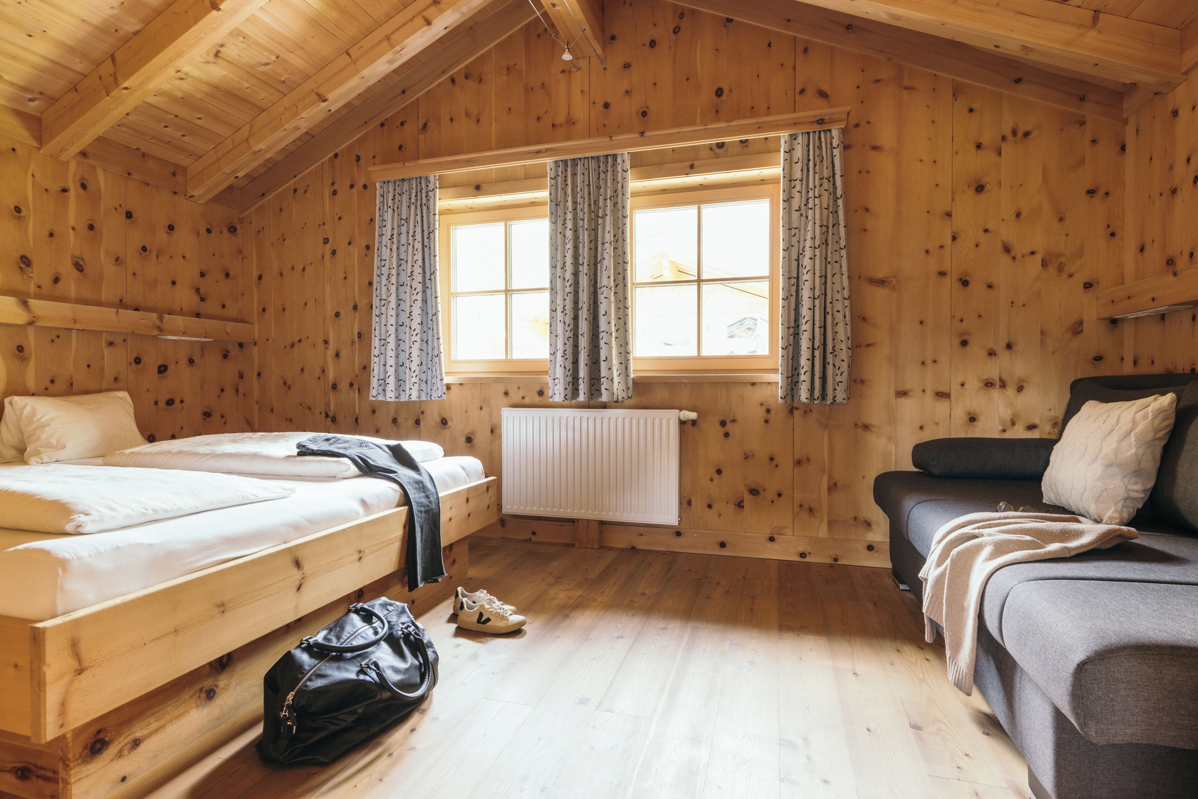 A wooden cabin room with two beds, a sofa, a radiator, and a window with curtains.