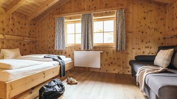 A wooden cabin room with two beds, a sofa, a radiator, and a window with curtains.