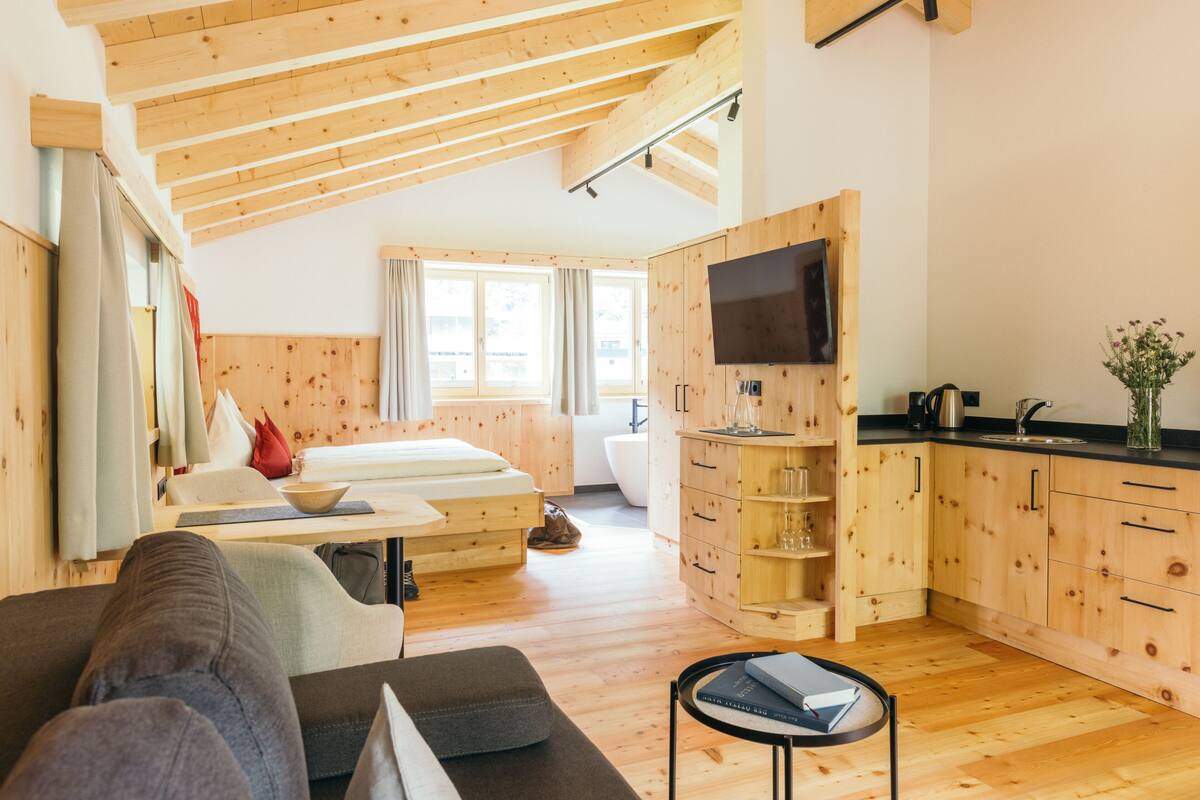 Een moderne, goed verlichte kamer met houten plafonds en vloeren, een bed, een bank, een televisie en een keukenhoek.