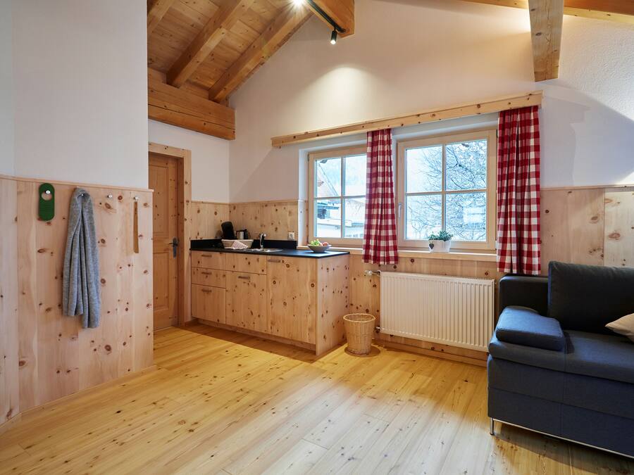 Una habitación acogedora con paredes y suelos de madera, una zona de cocina con mostrador y armarios, una ventana con cortinas de cuadros rojos y blancos, y un sofá de color oscuro.