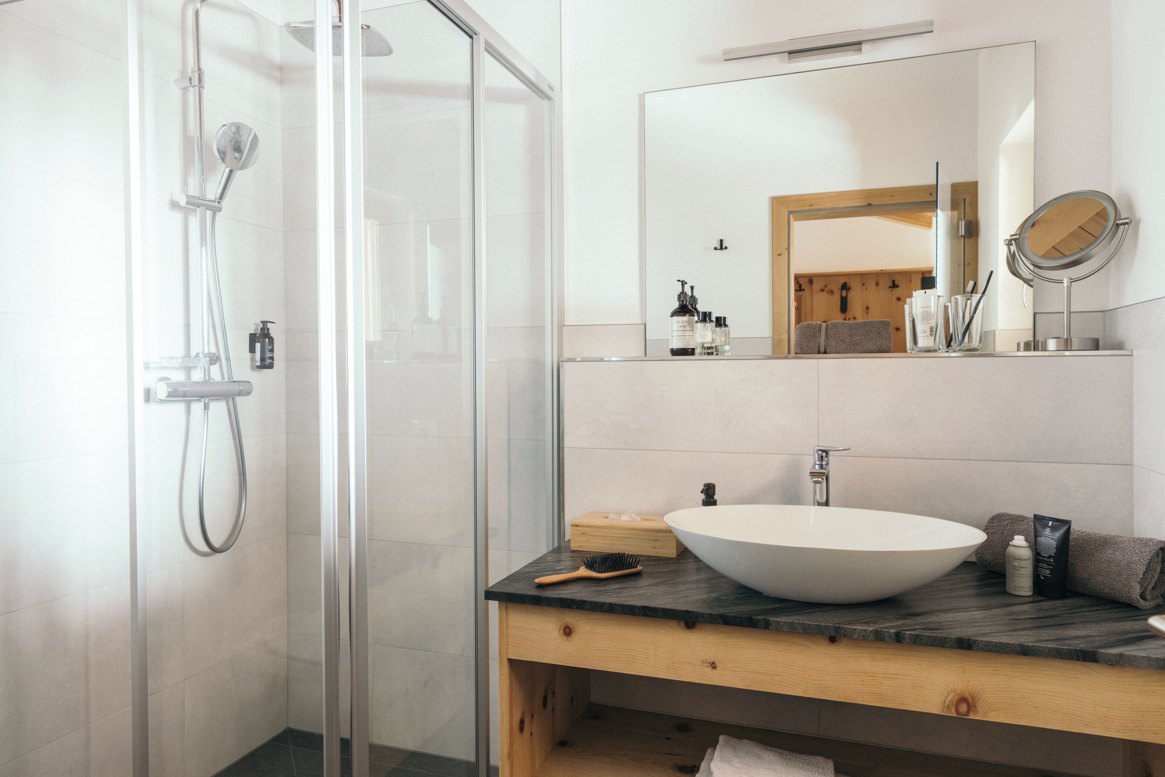 Moderni kylpyhuone, jossa on lasiseinällä varustettu suihku, suuri pesuallas sekä taso, jossa on peili ja hygieniatarvikkeita.