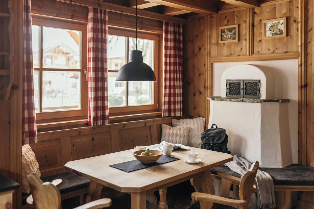 Een gezellig houten blokhut interieur met een eettafel, stoelen, een bankje, een hangende hanglamp en een raam met gordijnen.