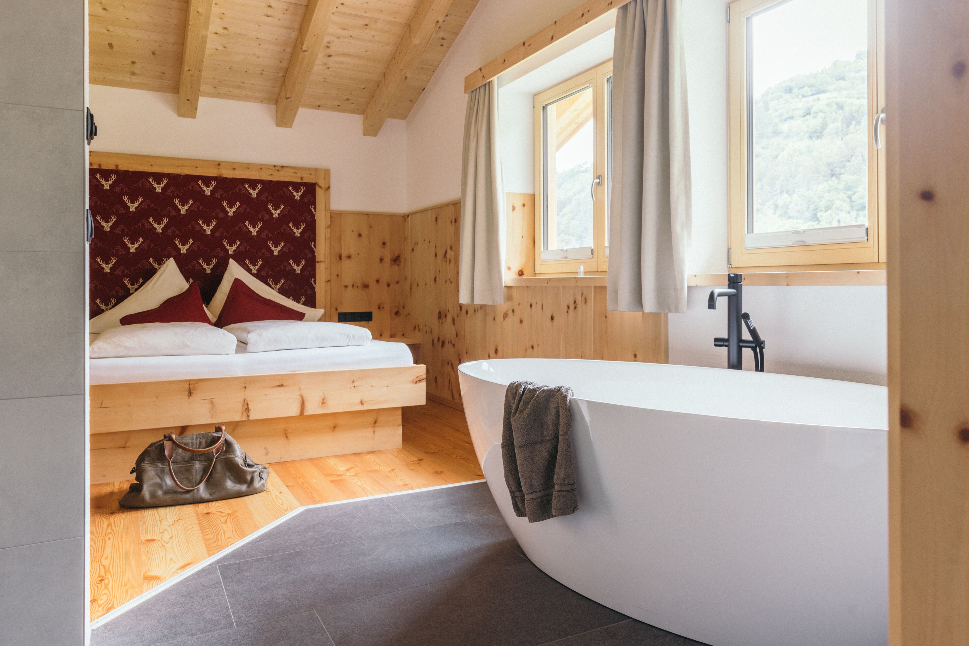 Un dormitorio moderno con techo de madera, una bañera grande, una cama con ropa de cama roja y blanca, y vista a un área boscosa a través de una ventana.