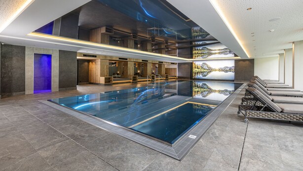 A modern indoor swimming pool with a clear view of the sky through a large glass ceiling, surrounded by lounge areas and wooden paneling.