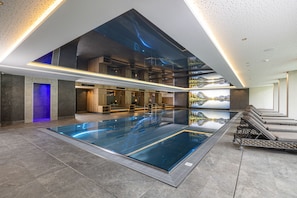 Una piscina interior moderna con vista despejada al cielo gracias a un amplio techo de vidrio, rodeada de zonas de descanso y paneles de madera.