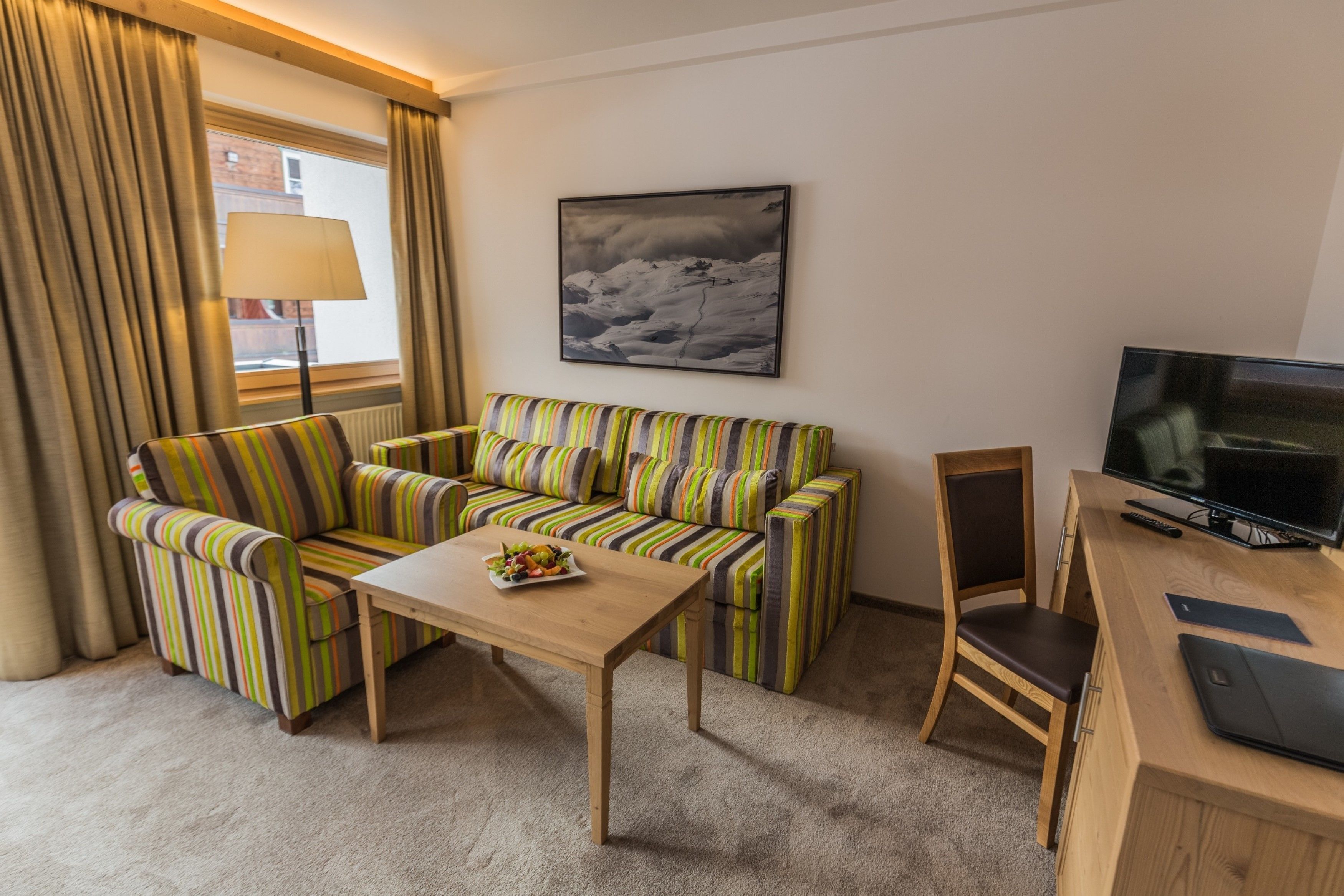 A hotel room with a striped sofa, a wooden table with a fruit bowl, a TV, and a lamp by the window.