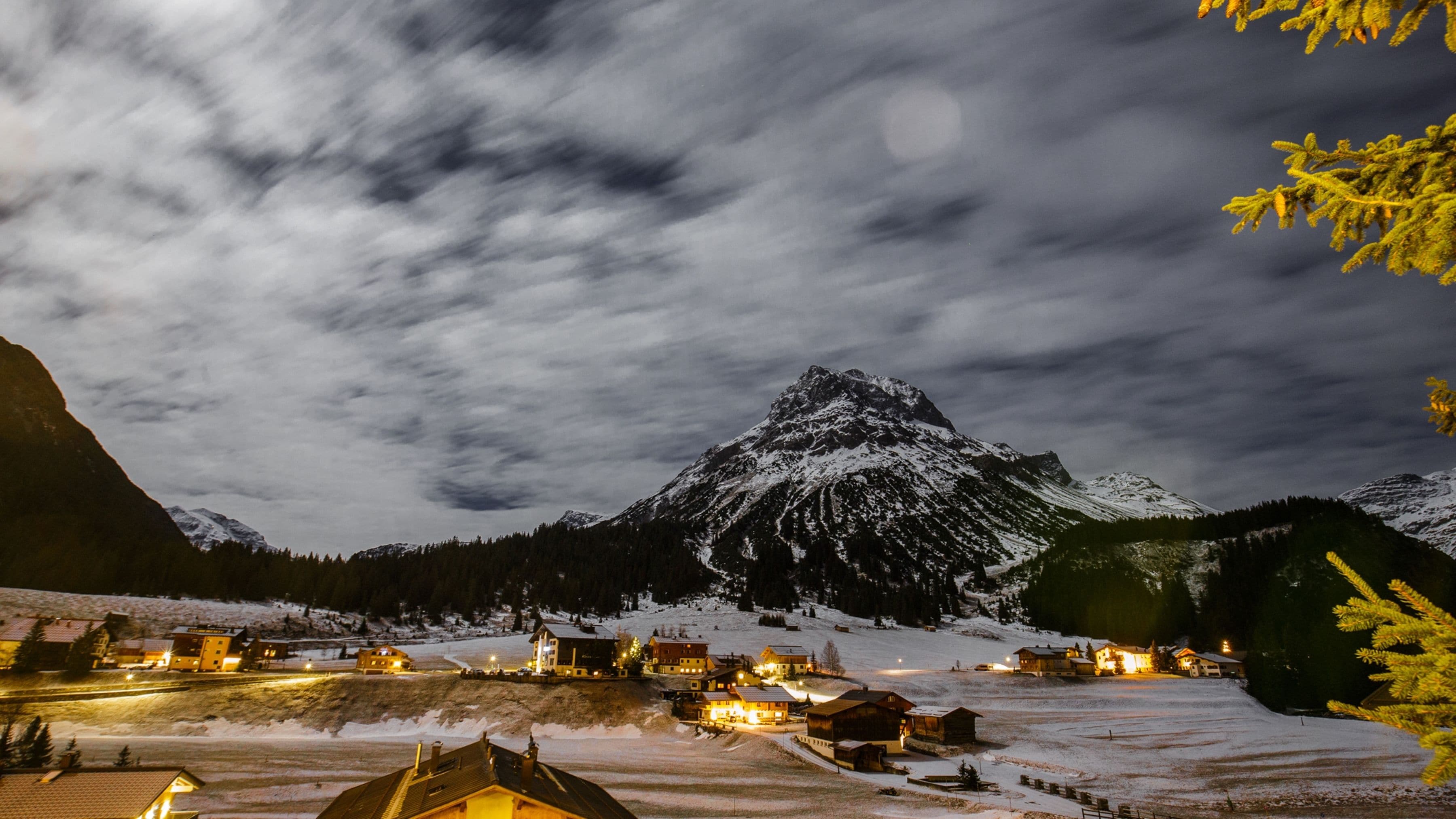 A snow-covered mountain village at night with illuminated buildings and a prominent peak.