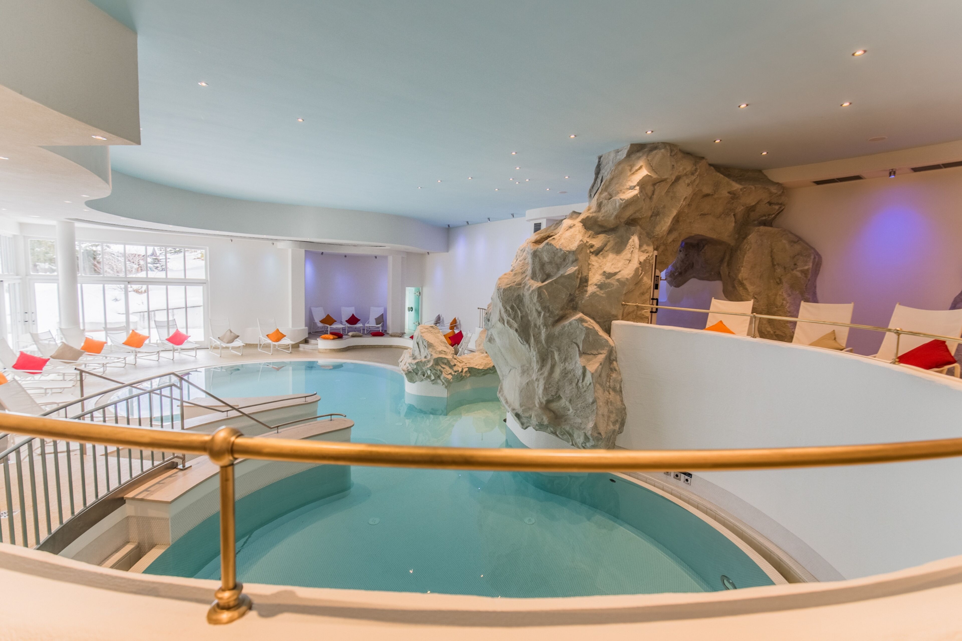 An indoor swimming pool with a rock feature, seating area with colorful cushions, and a staircase leading to a higher level.