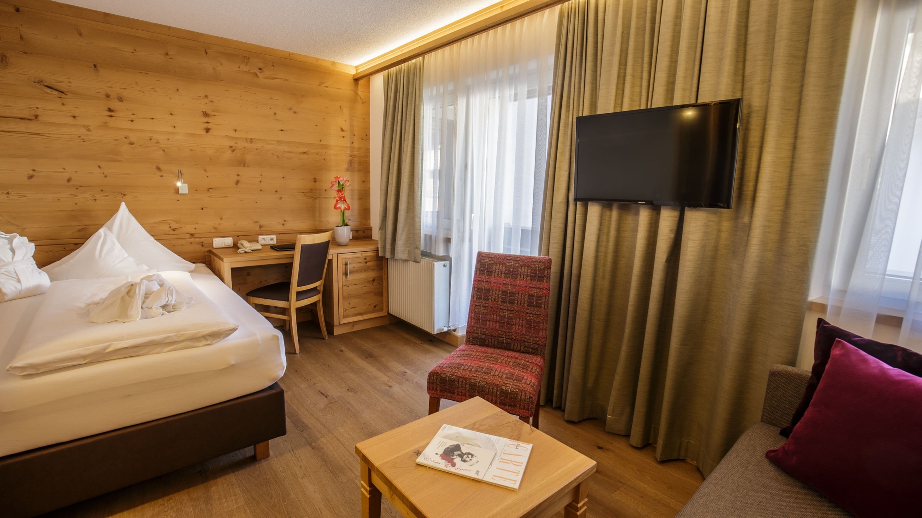 Habitación de hotel con pared de madera, una cama, un escritorio, una silla, un televisor y una ventana con cortinas.