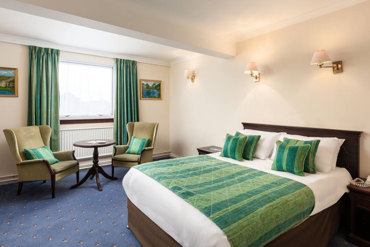 A hotel room with a large bed, two armchairs, a small table, and a window with curtains.