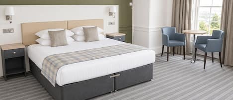 A hotel room with a large bed, two bedside tables, a small round table, two chairs, and a window with curtains.