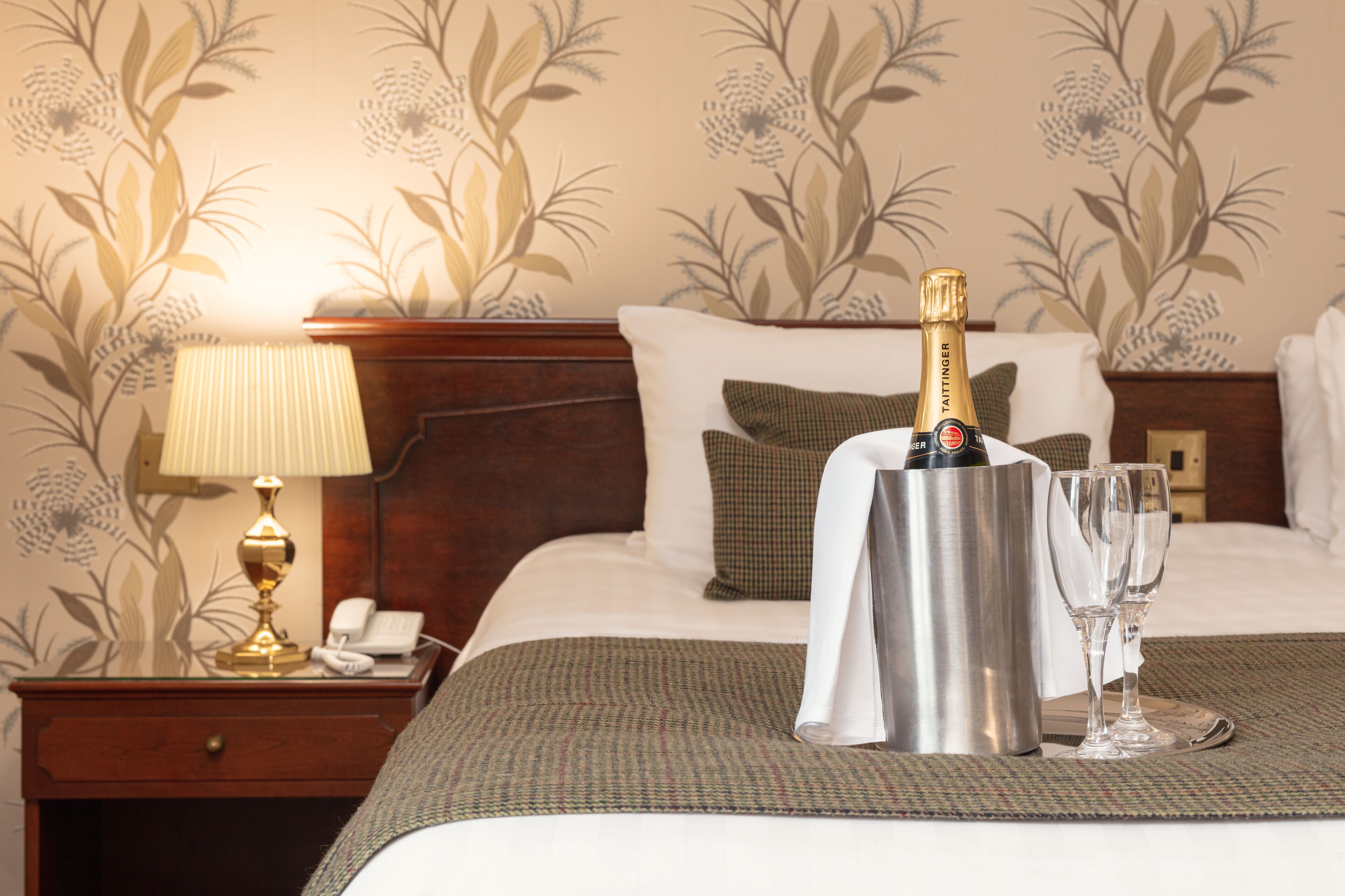 A hotel room with a bed, a nightstand with a lamp, a minibar, and a bottle of champagne with glasses.