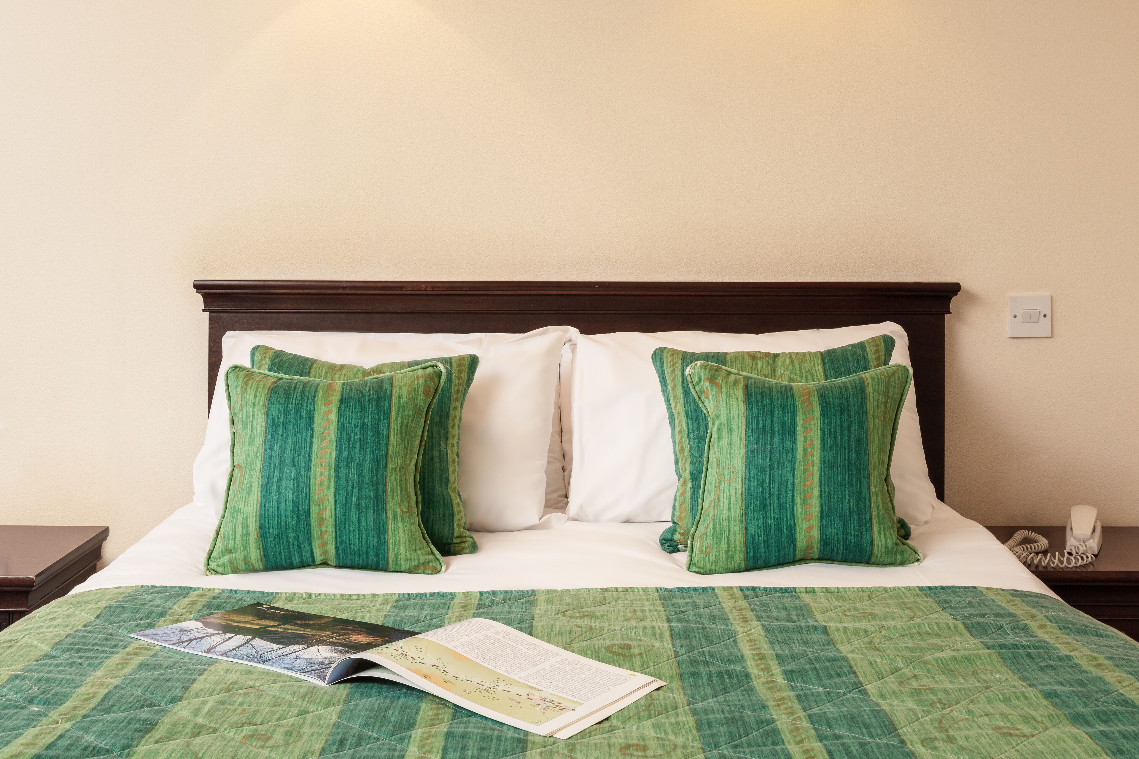 A neatly made bed with green and white bedding, a magazine on the bed, and a telephone on the nightstand.