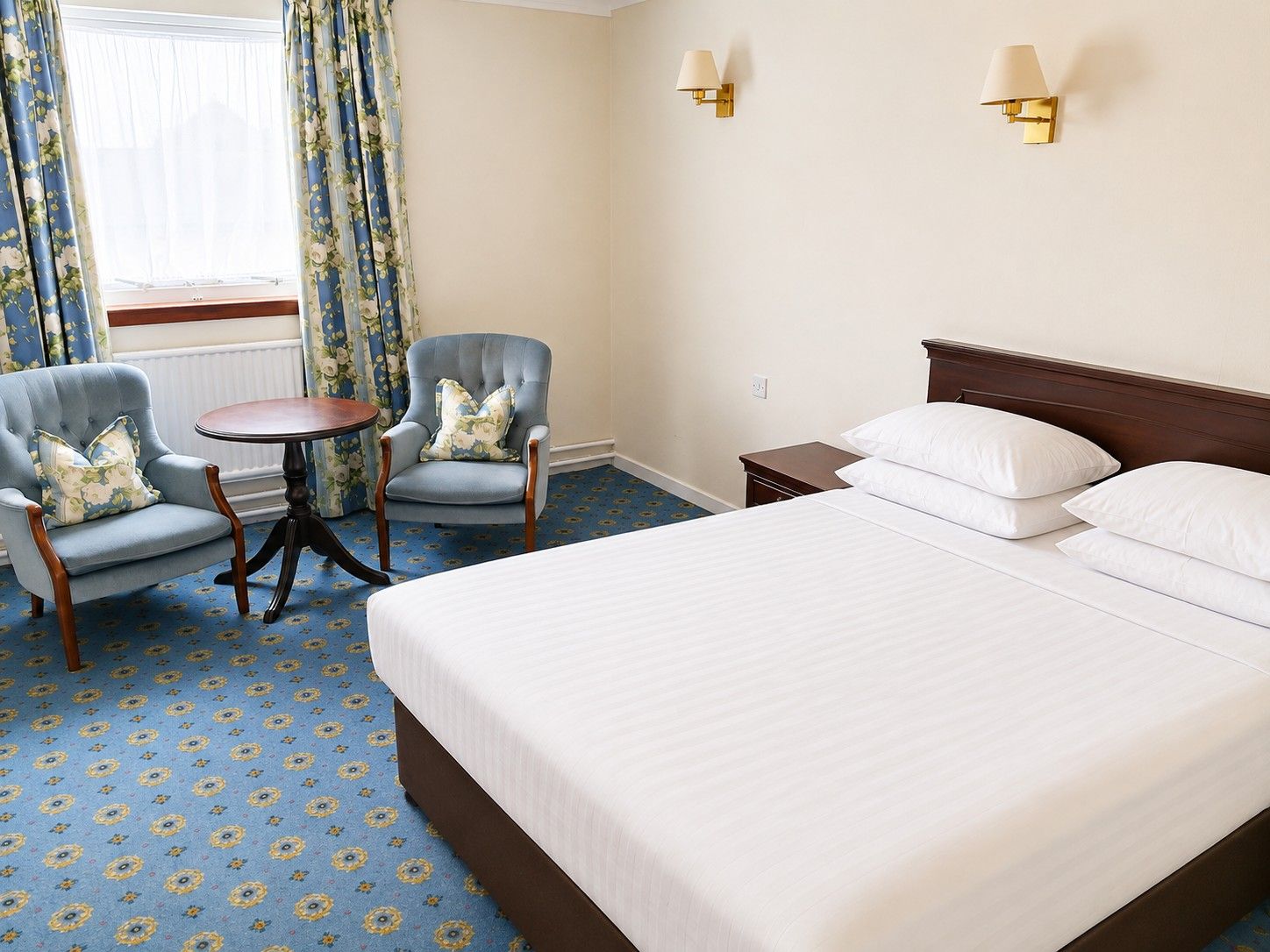 A hotel room with a large bed, two armchairs, a small table, and two wall-mounted lamps.