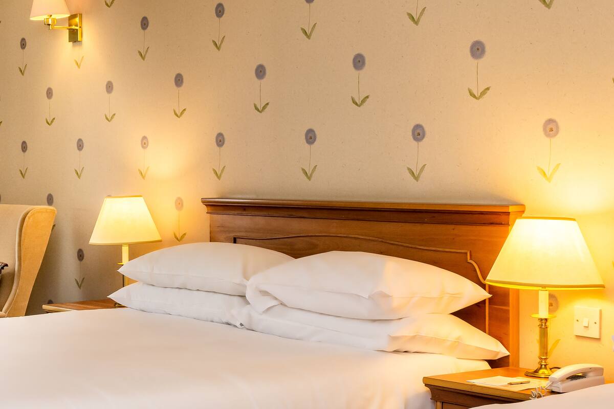 A hotel room with a neatly made bed, two bedside lamps, a wooden headboard, and floral wallpaper.