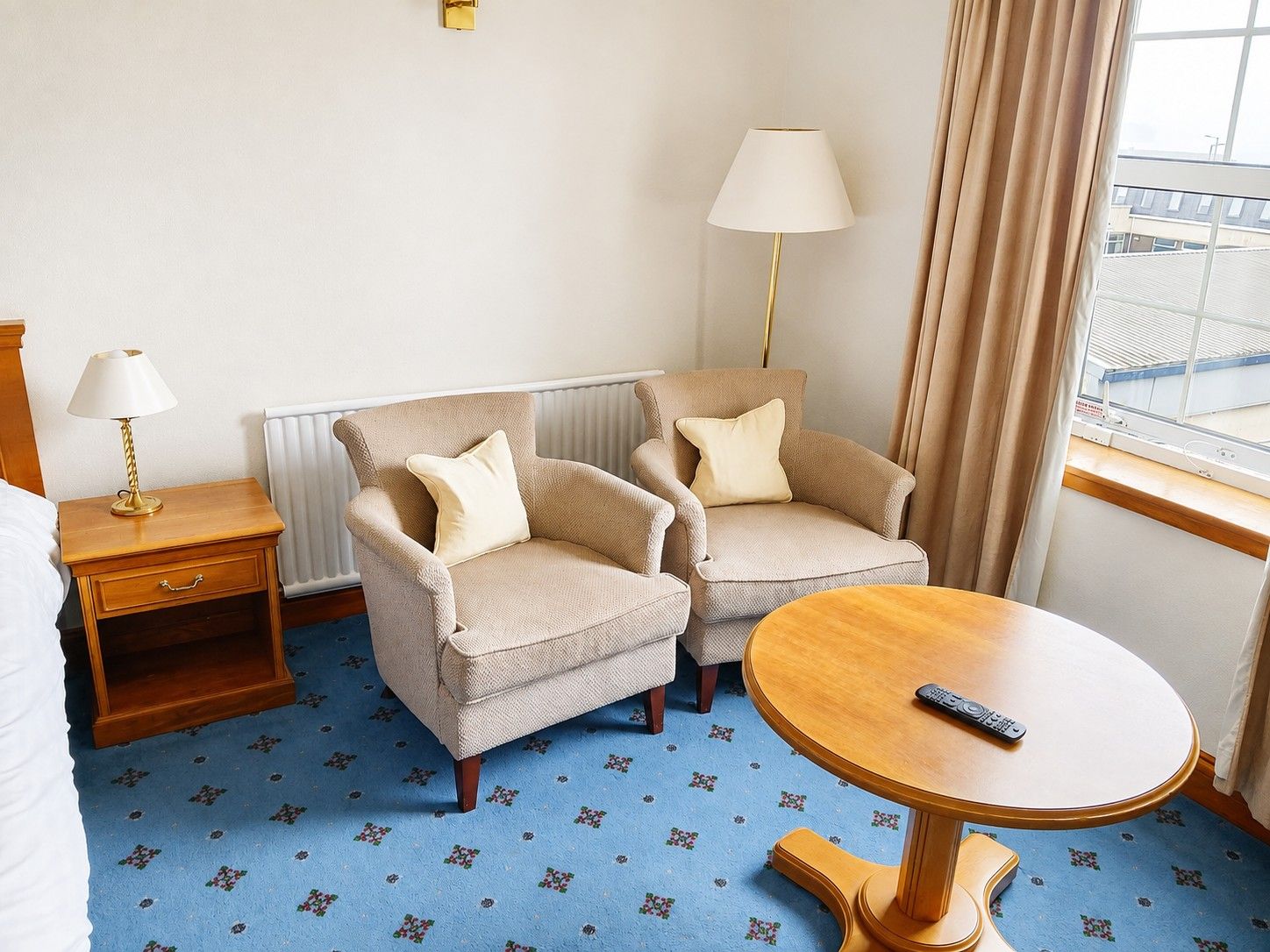 A hotel room with a bed, two armchairs, a small table, a lamp, and a view of the outside.