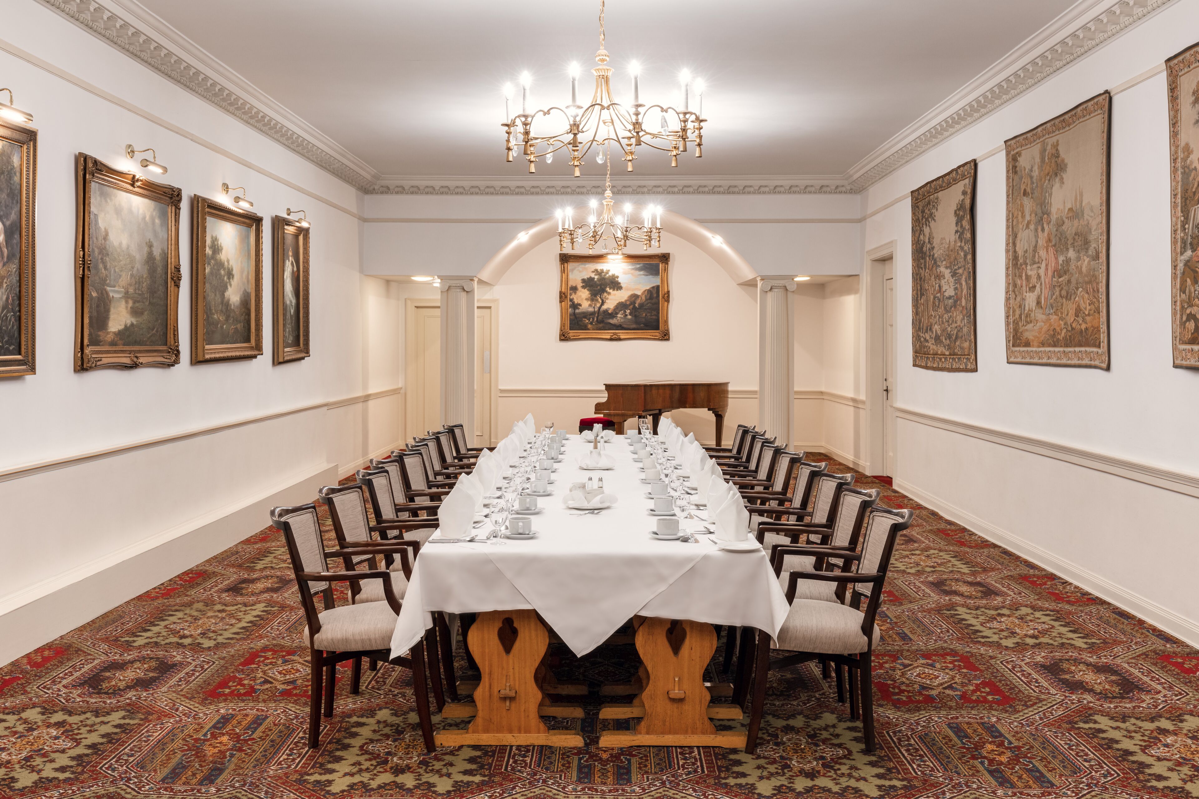 Une salle à manger élégante, avec une longue table dressée pour un banquet, entourée de chaises, et ornée de tableaux encadrés aux murs et d’un lustre au plafond.