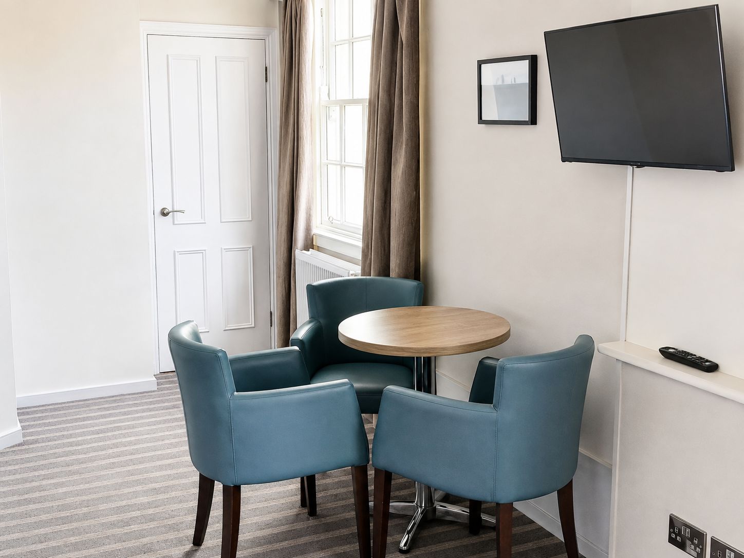 A hotel room with a round wooden table, two blue chairs, a flat-screen TV mounted on the wall, and a door.