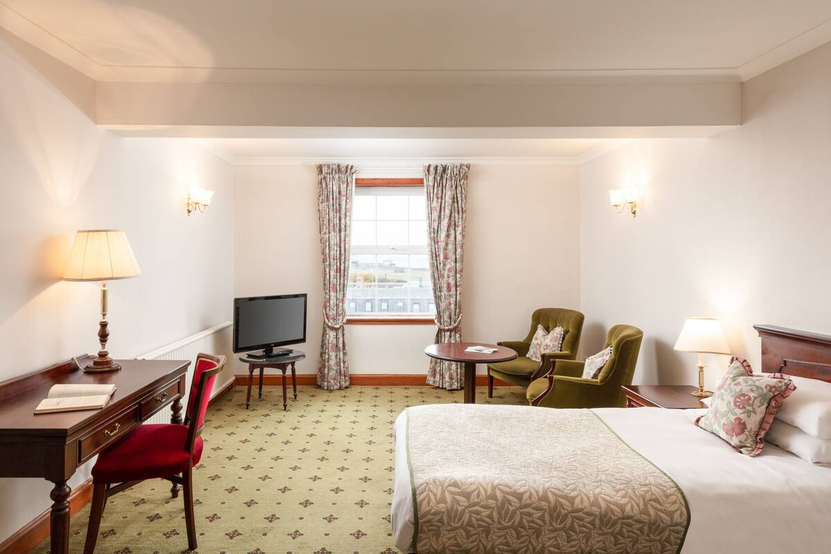A hotel room with a bed, a desk with a lamp, a television, two chairs, and a window with curtains.