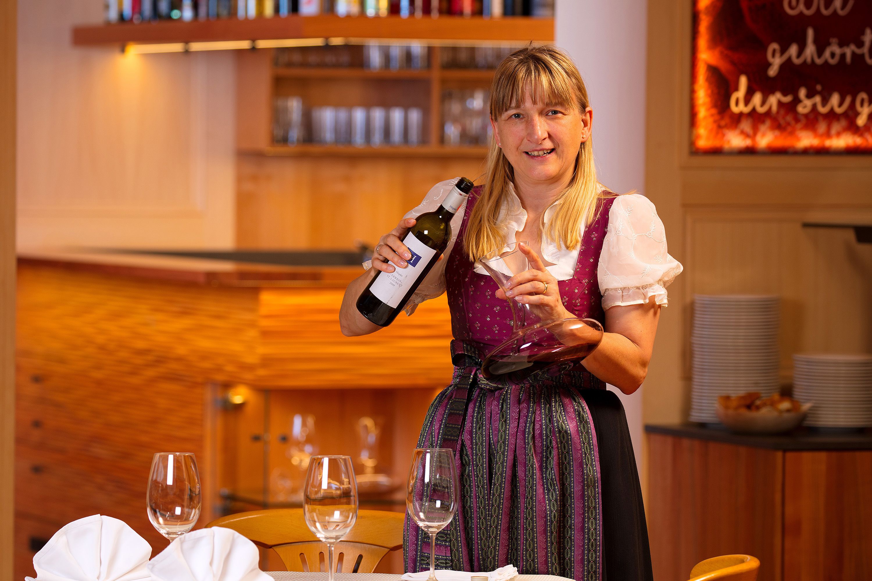 A person pouring wine into a glass at a table with several empty wine glasses.