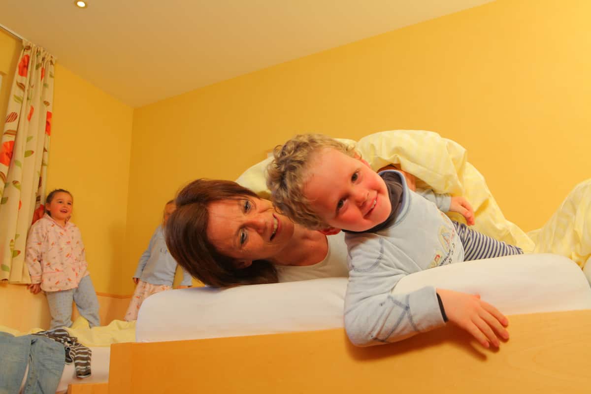 A room with a bed, a child lying on it, and another child standing by the window.