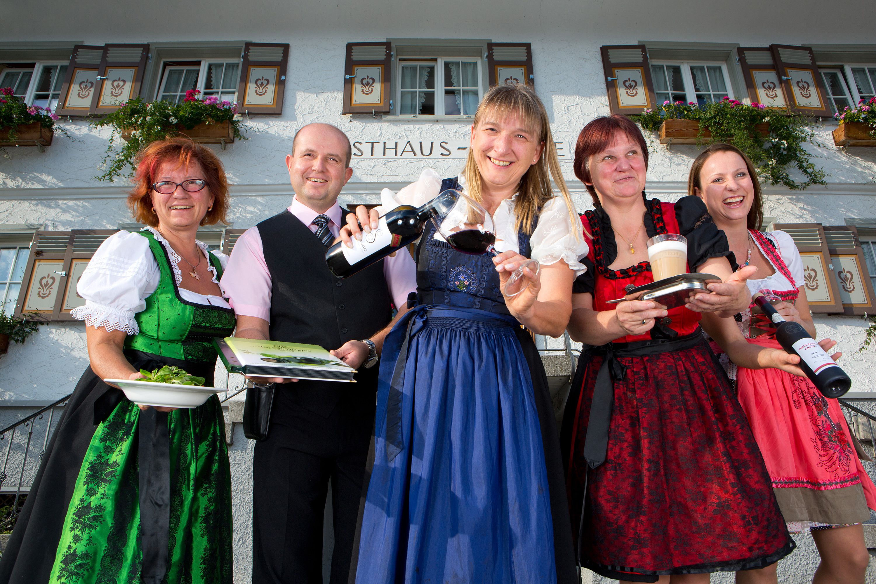 Un groupe de personnes, vêtues de costumes traditionnels, se tient devant un bâtiment sur lequel on peut lire l’inscription « STHAUS ».