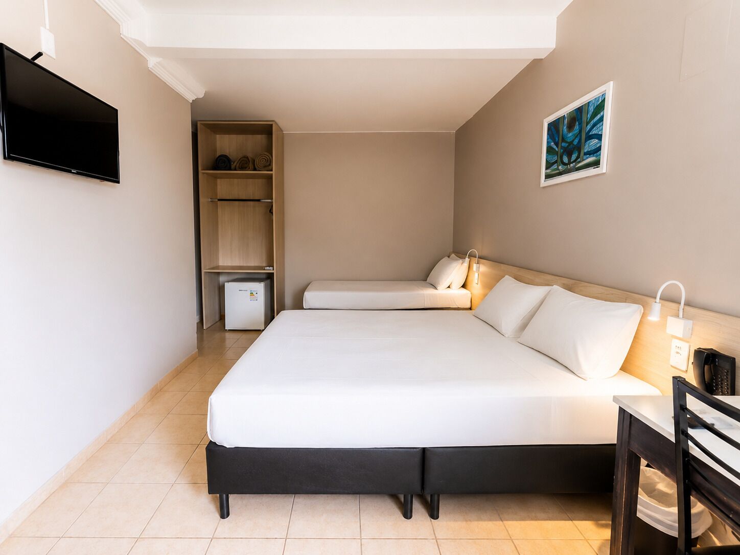 A hotel room with a large bed, a TV, a small fridge, a desk, and a painting on the wall.
