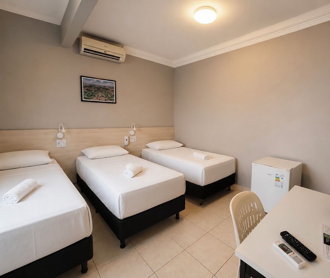 A hotel room with three single beds, a small table with a chair, a refrigerator, and an air conditioning unit.