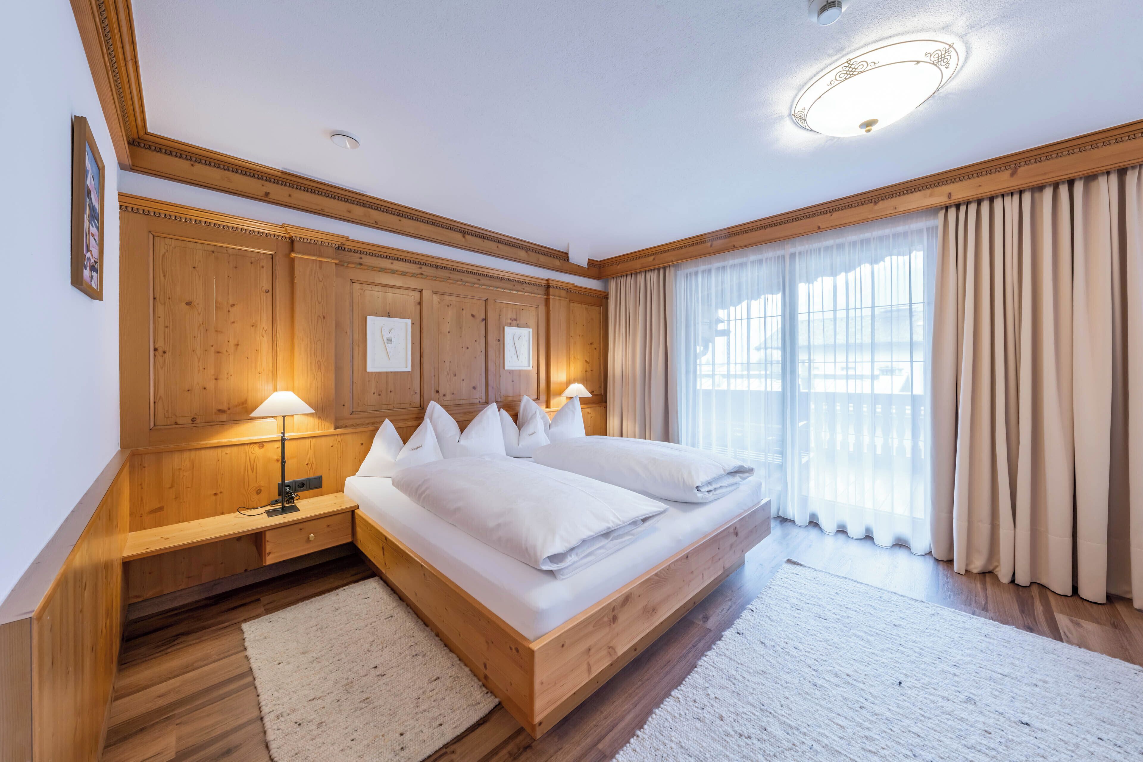 A bedroom with a wooden bed, bedside lamps, a nightstand, and a large window with curtains.