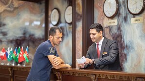 Deux hommes se tiennent derrière un comptoir d’accueil, avec des drapeaux et des horloges en arrière-plan.