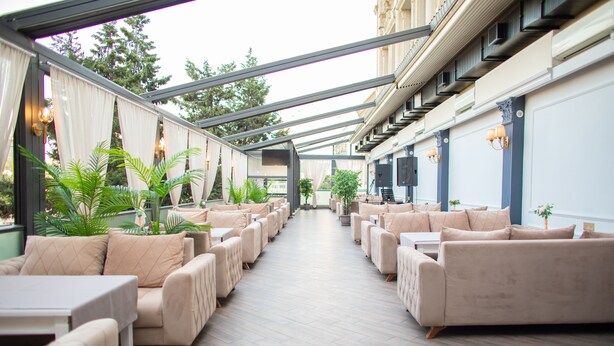 Un espace extérieur aménagé, abrité et doté de canapés et de tables de couleur beige, entouré de plantes en pot.