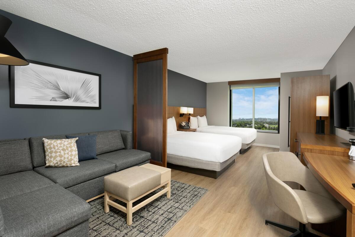 Een moderne hotelkamer met een grijze modulaire bank, een houten bureau, een bed met witte lakens en een groot raam met uitzicht op de stad.