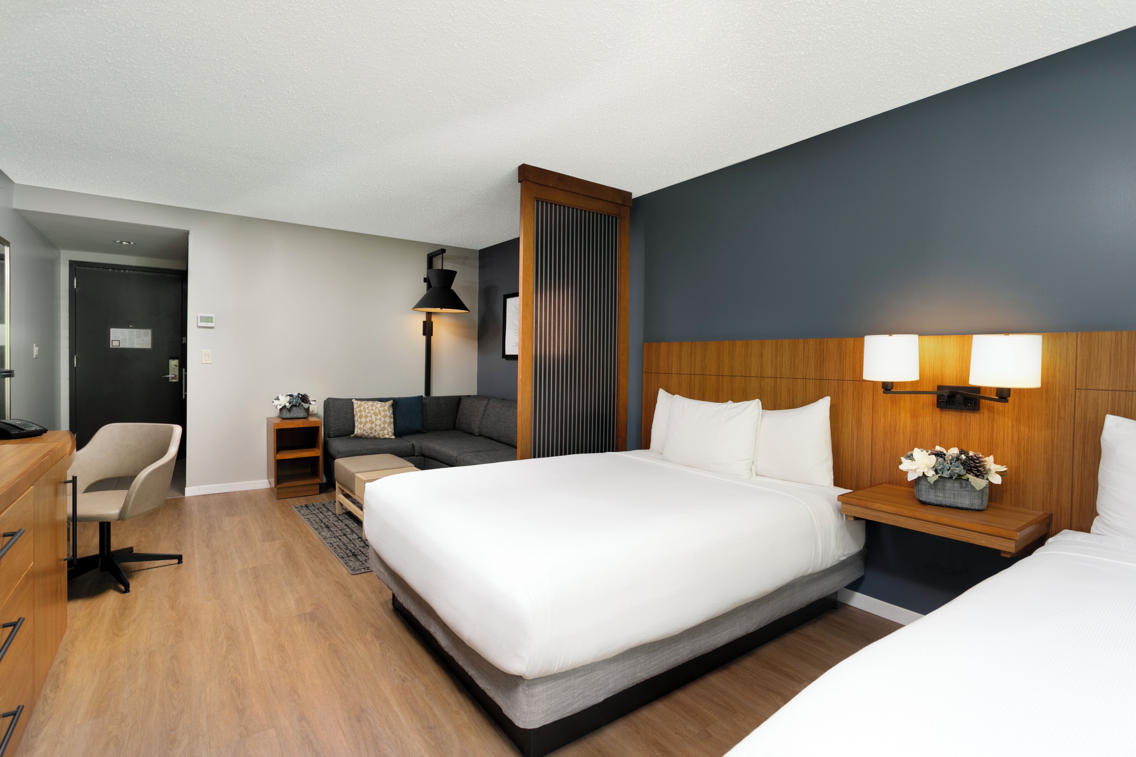 A hotel room with a large bed, a desk, a chair, a sofa, and a nightstand with a lamp.