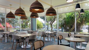 A modern indoor dining area with multiple tables and chairs, large windows with a view of greenery and a street, and pendant lighting. - Hyatt Place Bayamon (Bayamon)