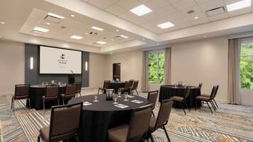 A conference room with multiple tables and chairs, a projector screen, and a large window with a view of greenery.