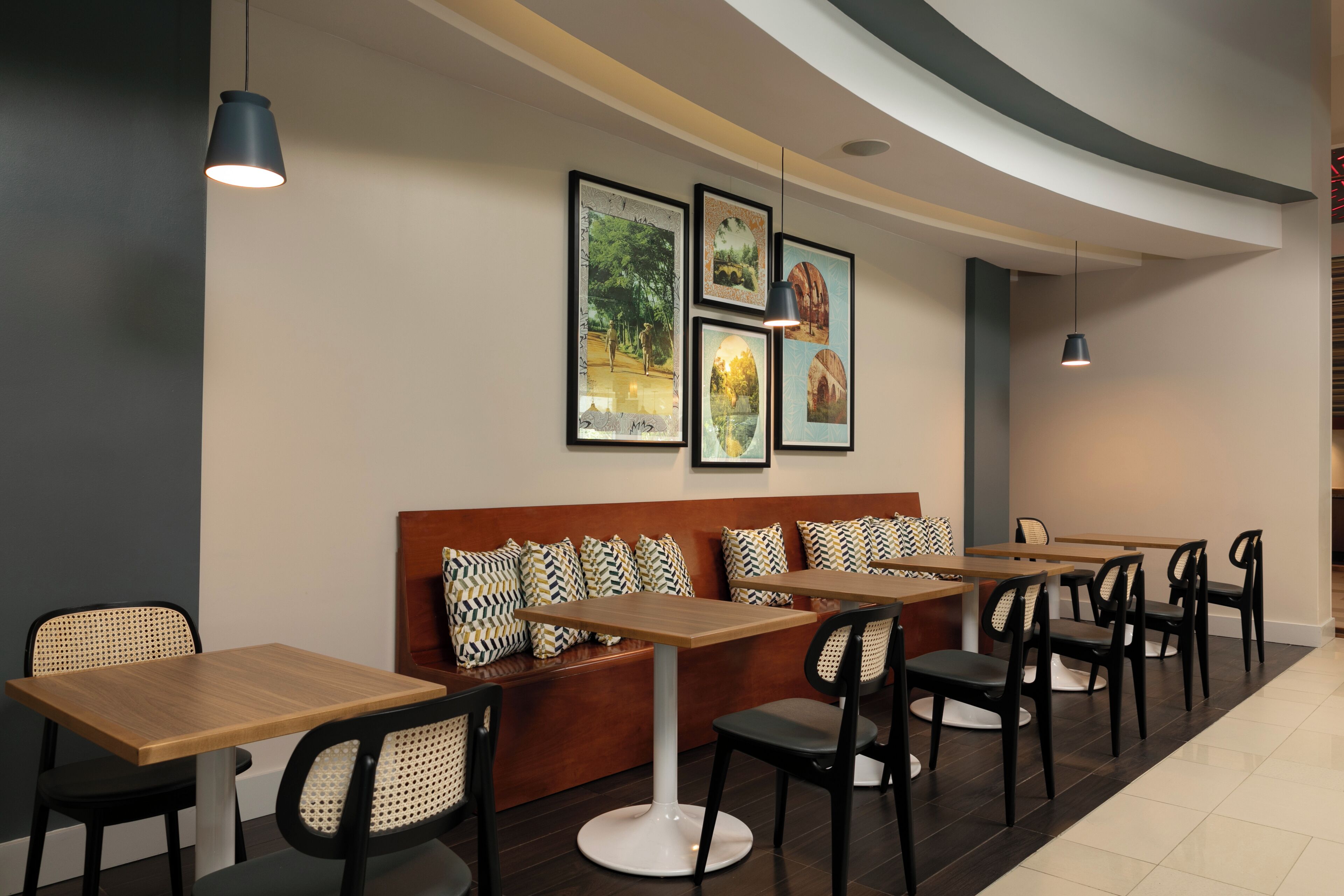A modern restaurant with wooden tables and black chairs, a brown sofa with patterned cushions, and framed pictures on the wall.