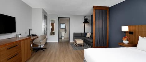 A hotel room with a wooden desk, a bed, a sofa, a TV, and a bathroom visible through an open door.