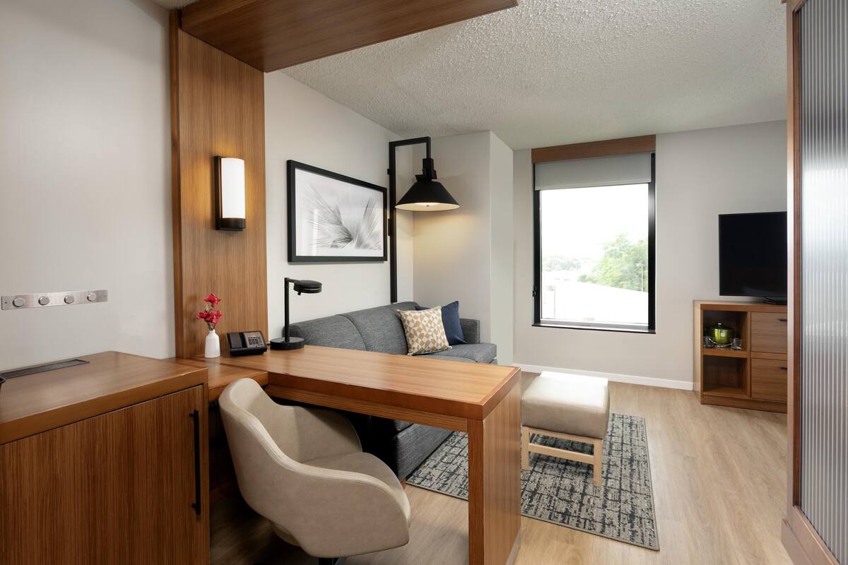 Een moderne woonkamer met een houten bureau, een grijze bank, een zwarte vloerlamp en een groot raam met uitzicht op groen.